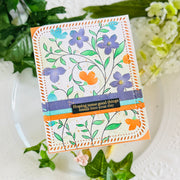 A handmade greeting card with a floral design, featuring purple flowers, green leaves, and orange accents, wrapped with a purple and orange ribbon and a black banner that reads 'Hoping some good things sneak into your day'. The card is displayed on a white plate with green leaves and white flowers in the background.