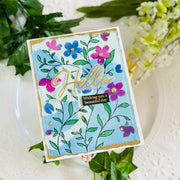 A handmade greeting card with a light blue background, decorated with hand-painted flowers in pink, purple, and blue, green leaves, gold foil script reading 'Hello', and a small black label that says 'Wishing you a beautiful day'. The card is styled on a white plate with green and white flowers.