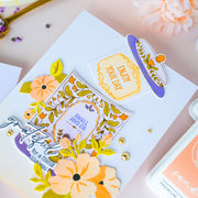 A close-up of a handmade layered card featuring floral elements, decorative text, and small gold embellishments on a pale pink background.