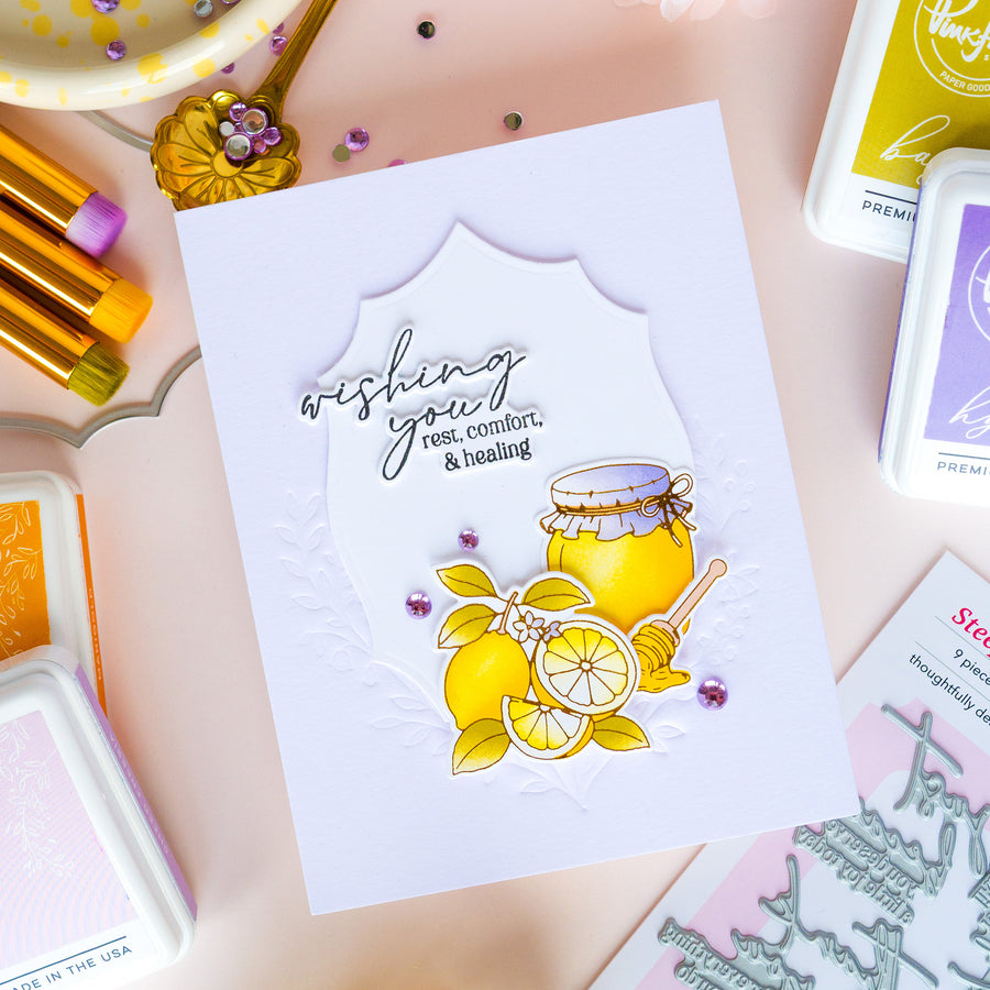 A handmade greeting card featuring a white scalloped background, silver embossed text reading 'wishing you rest, comfort, & healing,' and a yellow honey jar and lemon illustration with small purple rhinestone accents. The card is photographed on a pink surface with craft supplies and ink pads around it.