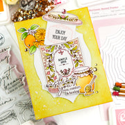 A handmade layered card with a yellow gradient background, featuring floral and fruit illustrations, a honeycomb motif, and layered white paper elements with decorative text. The card is styled on a flat-lay desk with other crafting tools and papers.