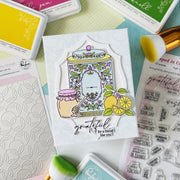 A handmade greeting card featuring a whimsical jar of honey illustration with lemons and the text 'grateful for a friend like you', styled in a flat-lay arrangement with craft supplies including ink pads, brushes, and a stamp sheet.