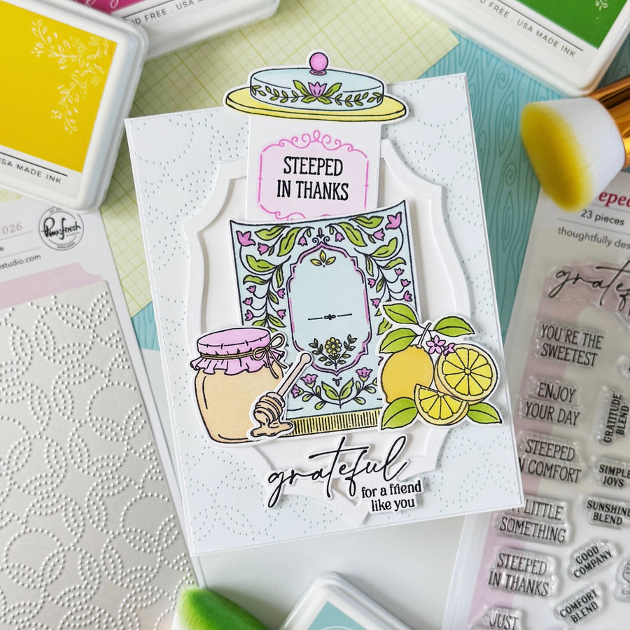 A handmade layered card featuring floral and fruit illustrations, with the phrases 'STEEPED IN THANKS' and 'grateful for a friend like you,' surrounded by craft supplies including ink pads, a die-cut sheet, and adhesive.