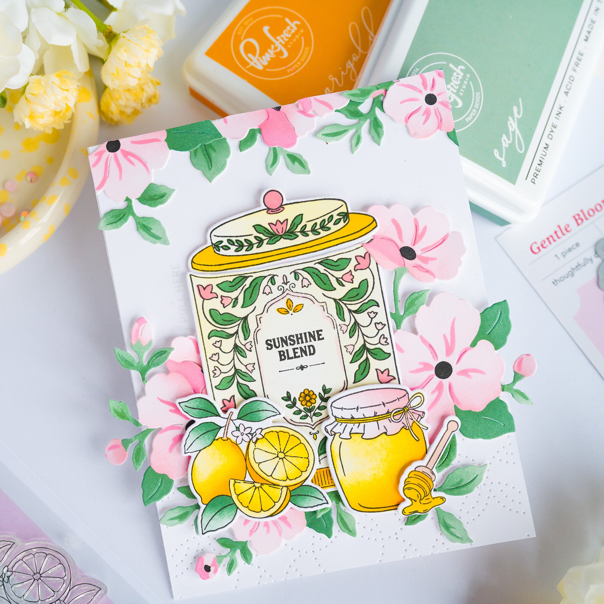 A handmade greeting card featuring a colorful illustration of honey jars, lemons, and pink flowers with the text 'SUNSHINE BLEND'. The card is styled on a white surface with crafting tools and flowers in the background.