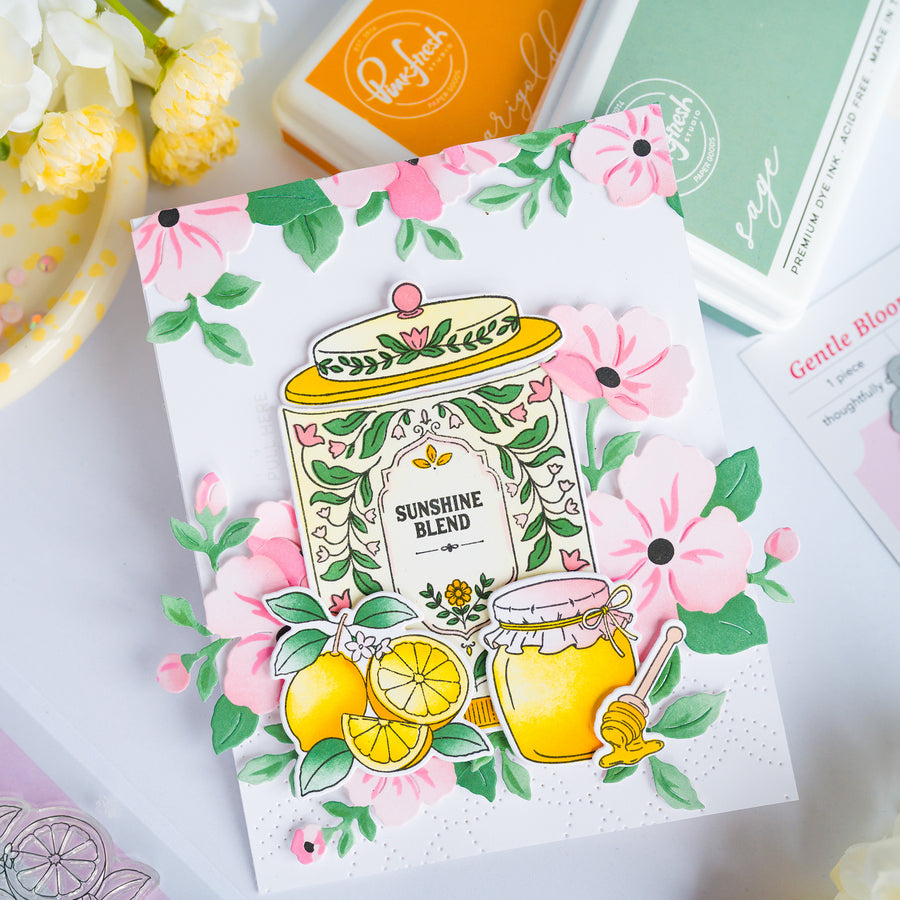 A handmade greeting card featuring a colorful illustration of honey jars, lemons, and pink flowers with the text 'SUNSHINE BLEND'. The card is styled on a white surface with crafting tools and flowers in the background.