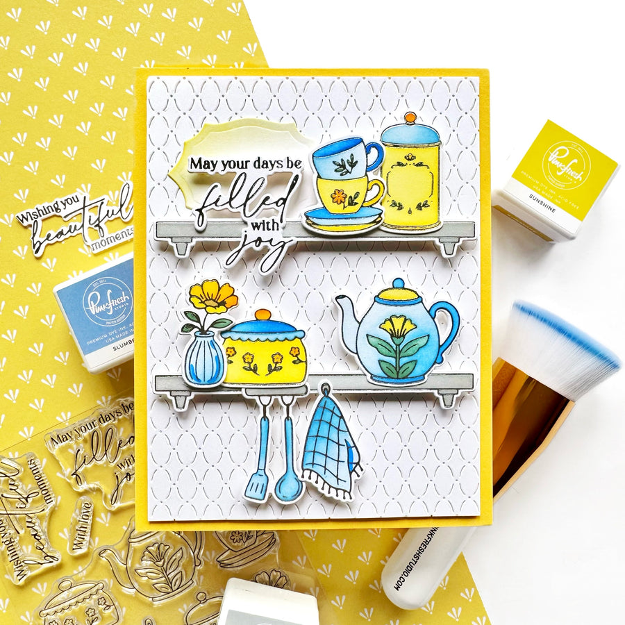 A handmade greeting card featuring a yellow border and a white patterned background with illustrated kitchen items on gray shelves, including a teapot, teacup, flowers, and utensils. The card is styled on a white surface with crafting supplies, including rubber stamps, an ink pad, and a paintbrush.