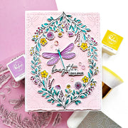 A handmade card featuring a colorful floral wreath and dragonfly design with the text 'brighter days ahead,' styled with crafting tools and stamps on a pink and white background.