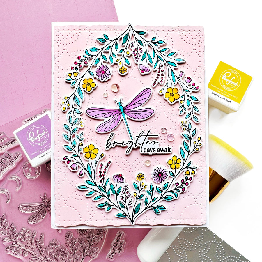 A handmade card featuring a colorful floral wreath and dragonfly design with the text 'brighter days ahead,' styled with crafting tools and stamps on a pink and white background.