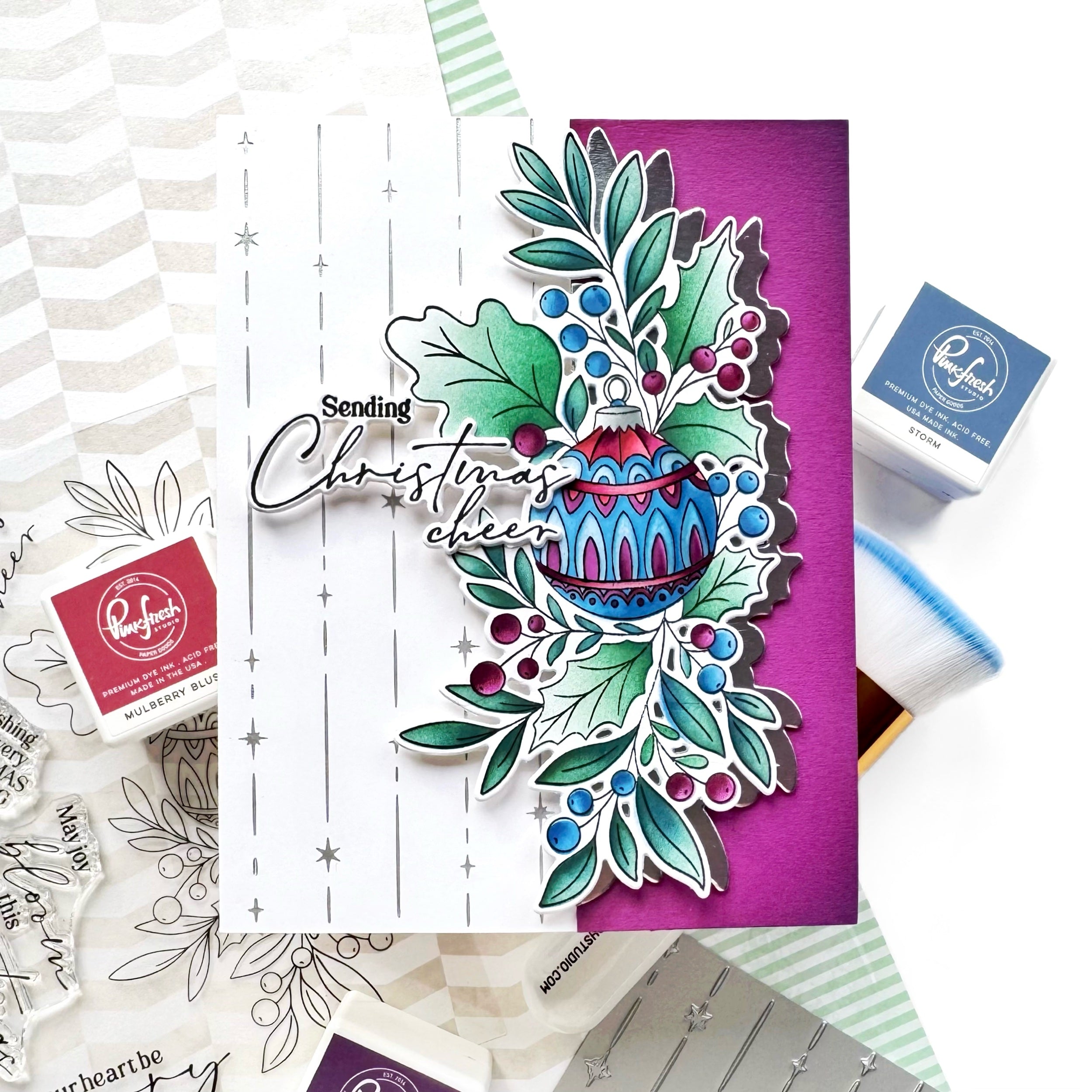 A handmade layered card featuring a Christmas ornament and holly design, styled with crafting supplies including Ranger ink pads and stamps on a white background.