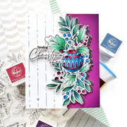 A handmade layered card featuring a Christmas ornament and holly design, styled with crafting supplies including Ranger ink pads and stamps on a white background.