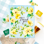 A handmade greeting card featuring yellow flowers and green leaves on a light blue background, with 'Thank you' written in black script. The card is styled in a flat-lay arrangement with crafting tools, stamps, and papers around it.