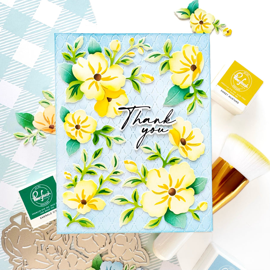 A handmade greeting card featuring yellow flowers and green leaves on a light blue background, with 'Thank you' written in black script. The card is styled in a flat-lay arrangement with crafting tools, stamps, and papers around it.