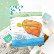 A handmade greeting card featuring an illustrated orange rowboat on blue water with the words 'HAPPY BIRTHDAY' at the top, styled with crafting supplies including stamps, an ink pad, and a paint brush on a white background.
