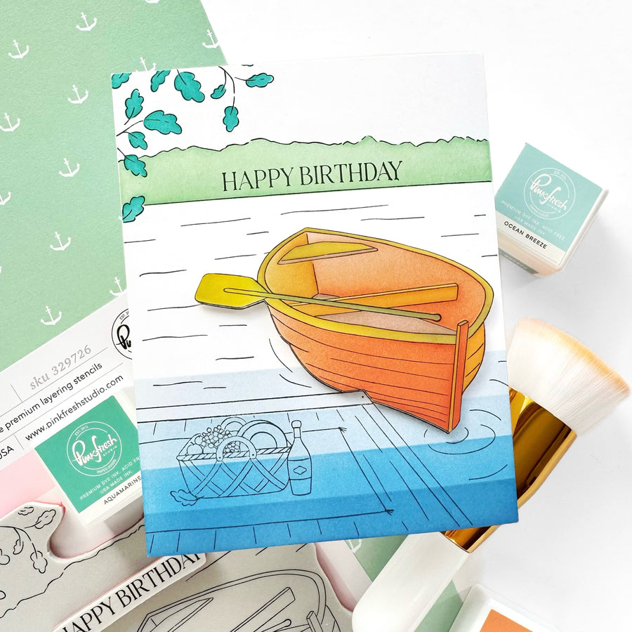 A handmade greeting card featuring an illustrated orange rowboat on blue water with the words 'HAPPY BIRTHDAY' at the top, styled with crafting supplies including stamps, an ink pad, and a paint brush on a white background.