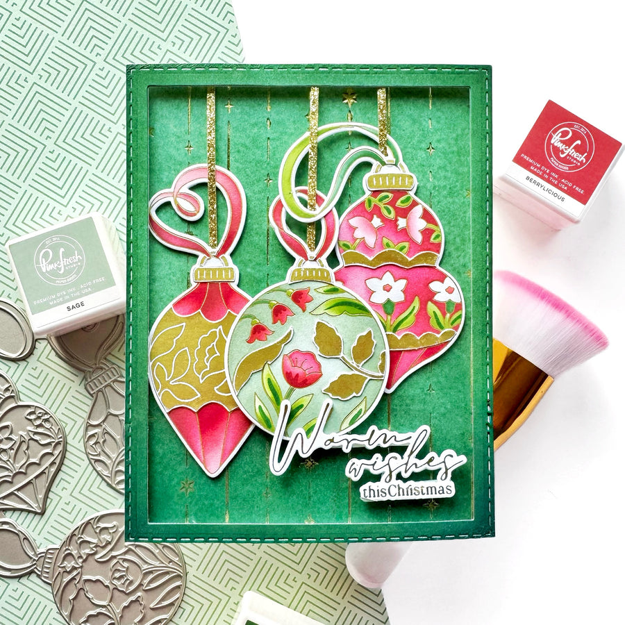 A handmade Christmas ornament card featuring three hanging ornament shapes in pink, green, and gold with floral and leaf patterns, displayed in a green frame. The card includes the text 'Warm wishes this Christmas' and is styled with Ranger stamps, ink pads, and a makeup brush on a white background.