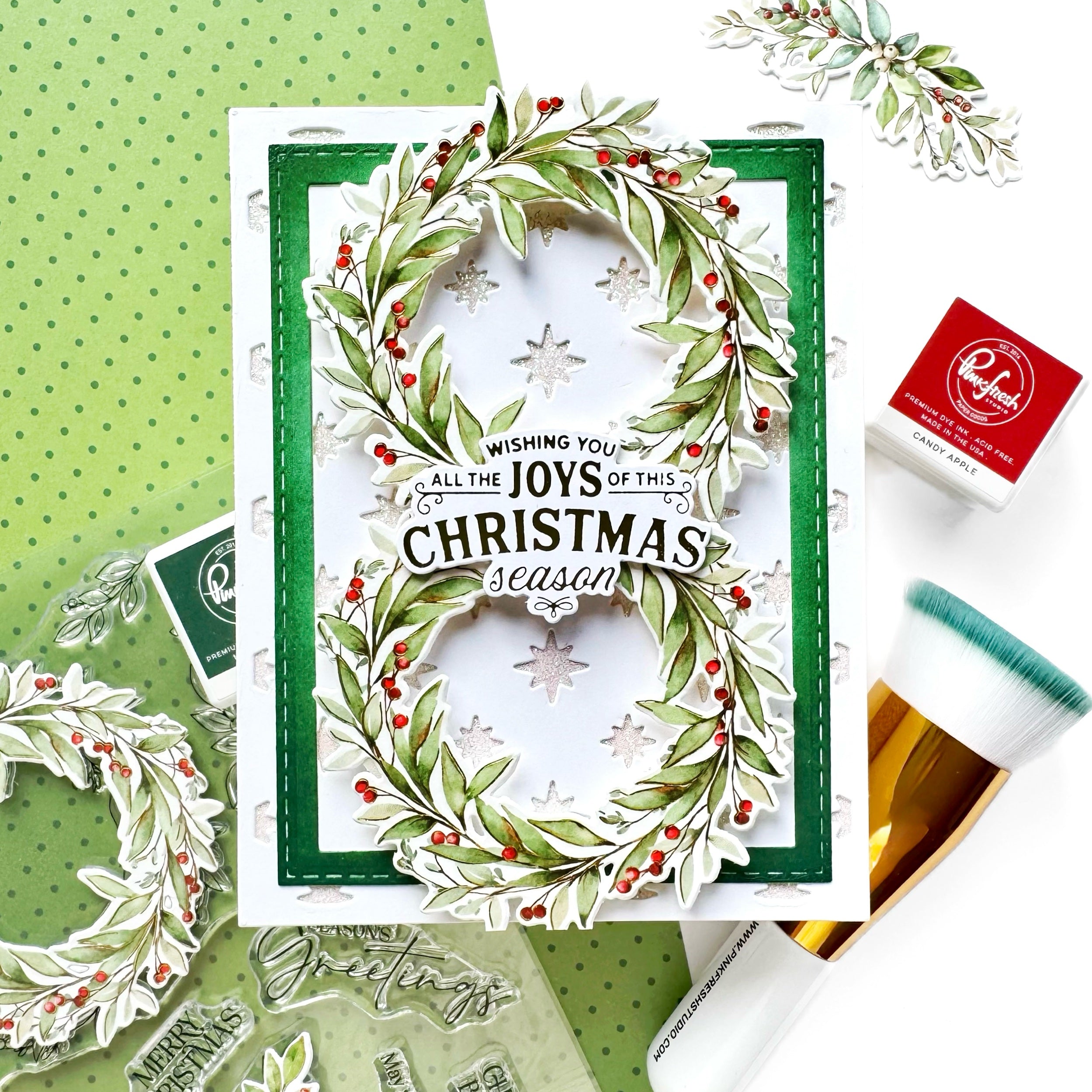 A handmade Christmas greeting card with a green border and a wreath design made from green leaves, white snowflakes, and red berries. The card features the text 'Wishing you all the JOYS of this CHRISTMAS season'.