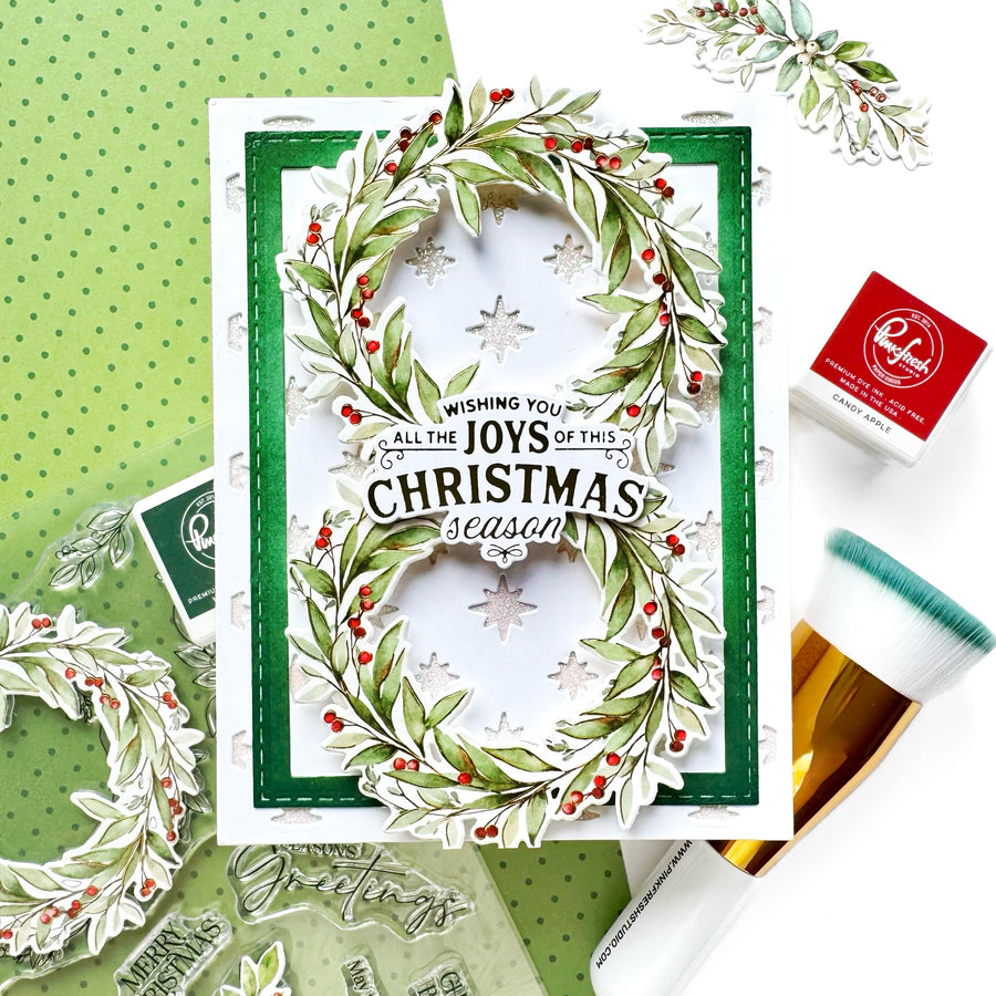 A handmade Christmas greeting card with a green border and a wreath design made from green leaves, white snowflakes, and red berries. The card features the text 'Wishing you all the JOYS of this CHRISTMAS season'.