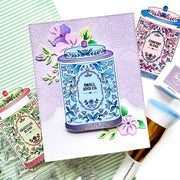 A flat-lay of handmade greeting cards featuring illustrated cosmetic jars with floral designs, alongside a makeup brush and stamping tools on a green striped background.