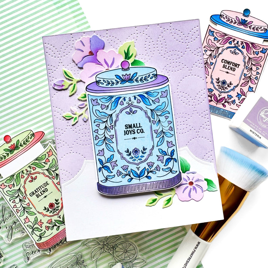 A flat-lay of handmade greeting cards featuring illustrated cosmetic jars with floral designs, alongside a makeup brush and stamping tools on a green striped background.