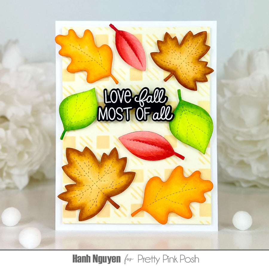 A handmade greeting card featuring autumn-themed die-cut leaves and the text 'LOVE fall MOST OF all' on a cream and yellow checkered background, displayed on a white surface with decorative white flowers in the background. The card is branded with 'Pretty Pink Posh' and designer 'Hanh Nguyen' at the bottom.
