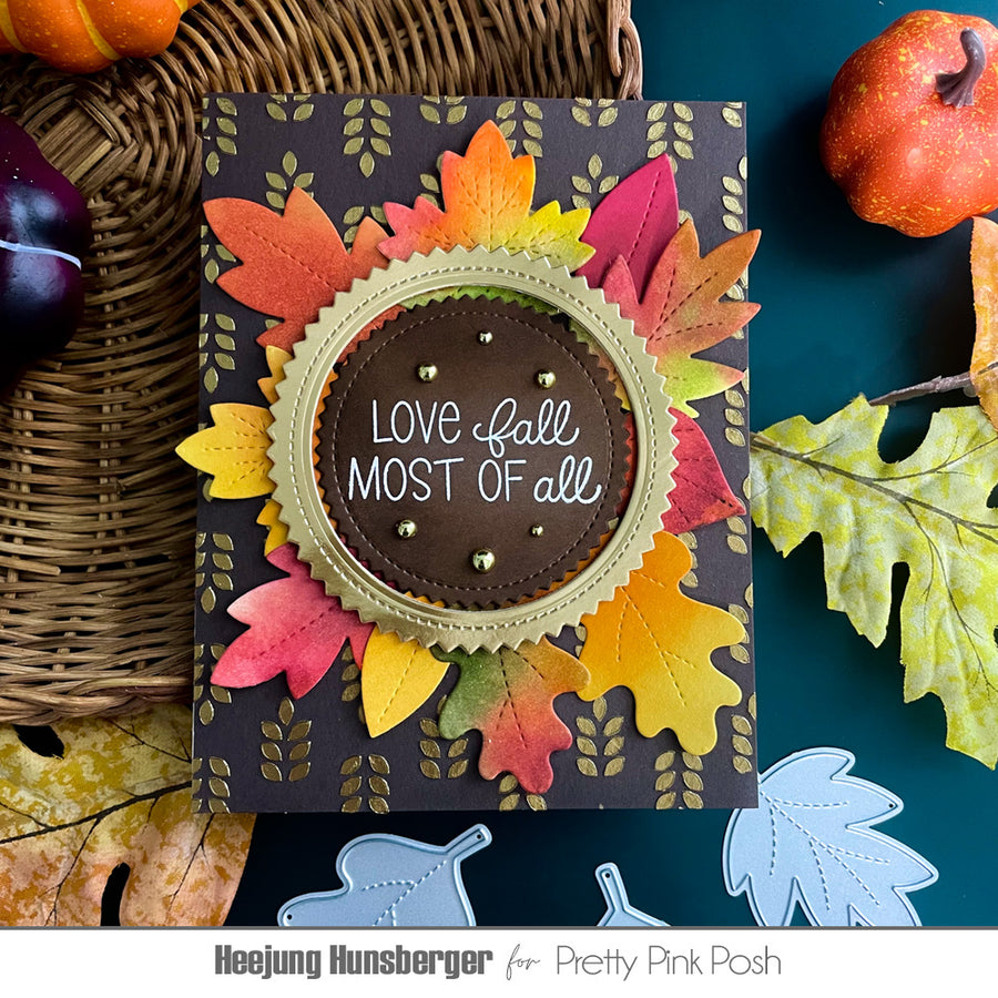A handmade autumn-themed greeting card featuring layered die-cut fall leaves in yellow, orange, pink, and green on a dark charcoal background. The center has a scalloped olive-green border around a brown circle with the phrase 'LOVE fall MOST OF all' in white script.