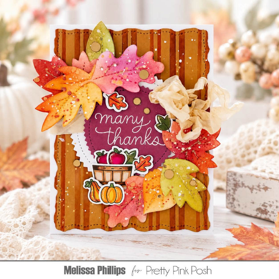 A handmade autumn-themed greeting card with layered paper leaves in orange, pink, yellow, and green, a purple center with the words 'many thanks,' and small fall illustrations including pumpkins and apples. The card is photographed on a soft, blurred rustic background with lace and fall leaves.