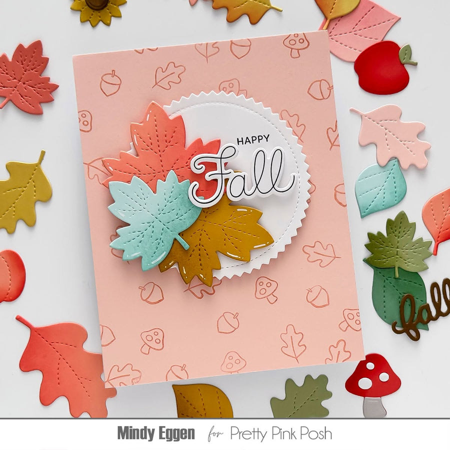 A handmade autumn-themed greeting card with layered die-cut fall leaves in coral, mint, and mustard on a pale pink patterned cardstock. The card has a scalloped white circle insert with the words “HAPPY Fall” in script.