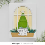 A handmade Christmas greeting card featuring a green tree silhouette framed by a white arched window, with a star on top and a banner reading 'Wishing You Warmth & Love' above greenery and red berries. The card is shown against a light gray background with decorative foliage in the corners.
