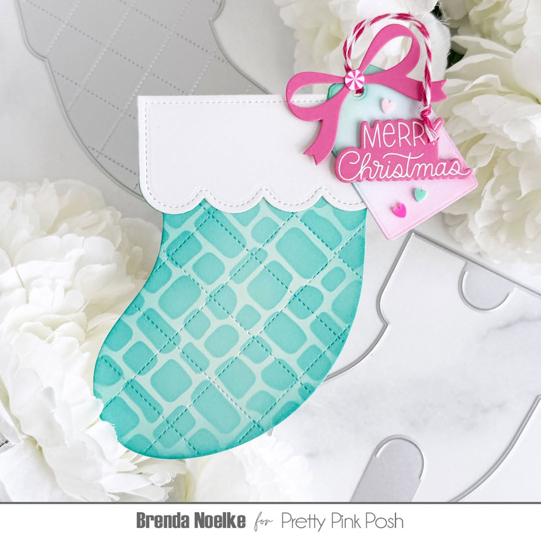 A Christmas stocking ornament or card embellishment in aqua and white, decorated with a pink ribbon and a hanging tag that reads “Merry Christmas,” styled among white flowers on a light surface.