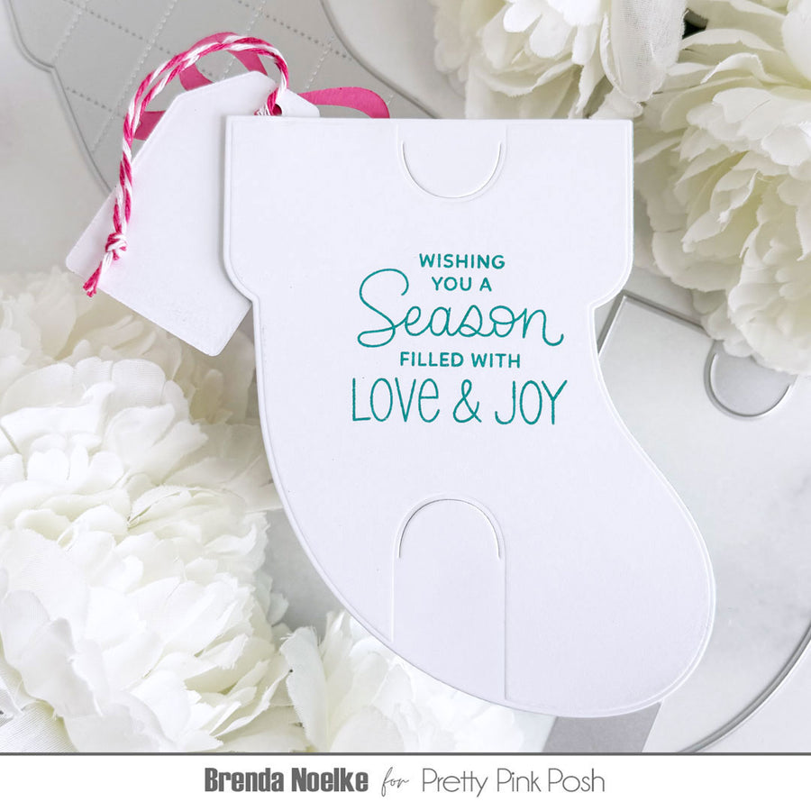 A white Christmas stocking-shaped paper ornament or card with teal text reading 'Wishing you a Season filled with Love & Joy,' hanging from a pink-and-white string, styled among white flowers on a light surface.