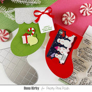 A styled flat-lay of Christmas stocking-shaped card-making or gift tags, including green and red stockings with glitter trim, candy canes, peppermint candies, a gift tag reading 'HAVE A HOLLY JOLLY CHRISTMAS,' and a clear stamp sheet. The layout is branded with 'Dana Kirby for Pretty Pink Posh' at the bottom.