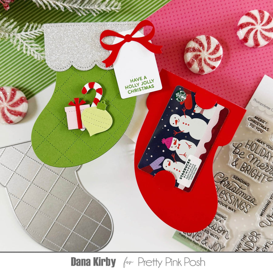 A styled flat-lay of Christmas stocking-shaped card-making or gift tags, including green and red stockings with glitter trim, candy canes, peppermint candies, a gift tag reading 'HAVE A HOLLY JOLLY CHRISTMAS,' and a clear stamp sheet. The layout is branded with 'Dana Kirby for Pretty Pink Posh' at the bottom.