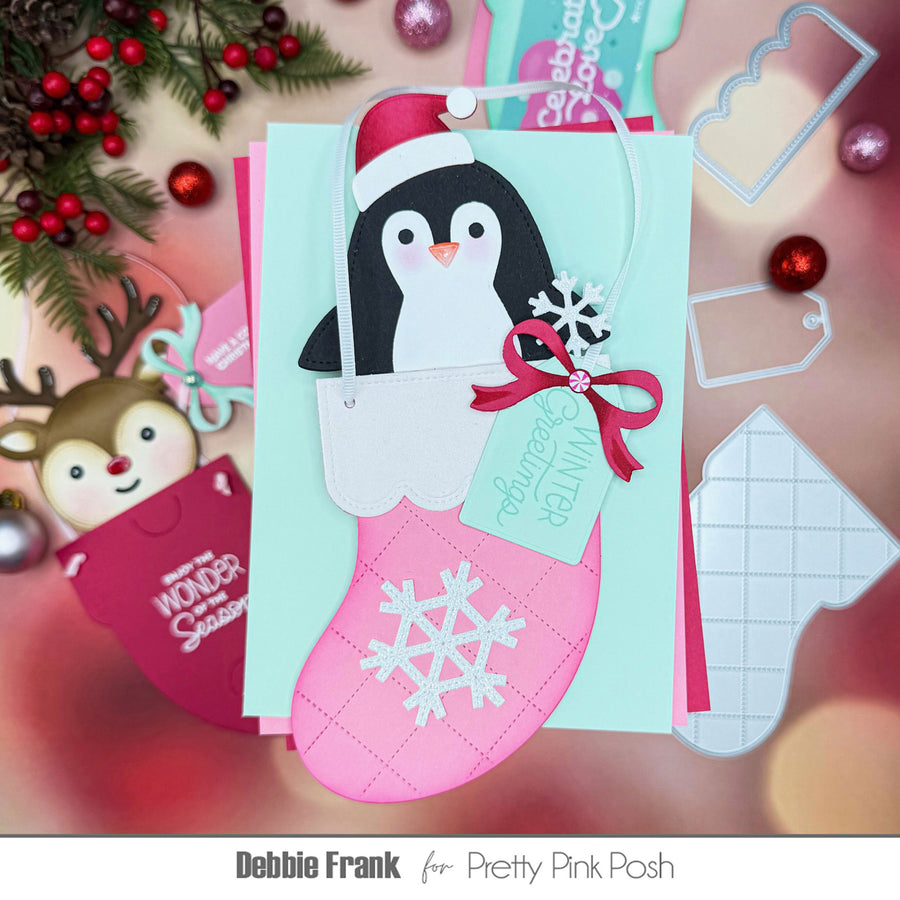 A festive handmade Christmas card or gift tag featuring a penguin in a pink stocking with a red Santa hat, red ribbon, and snowflake details, styled with holiday decorations and layered card elements on a soft pink background.
