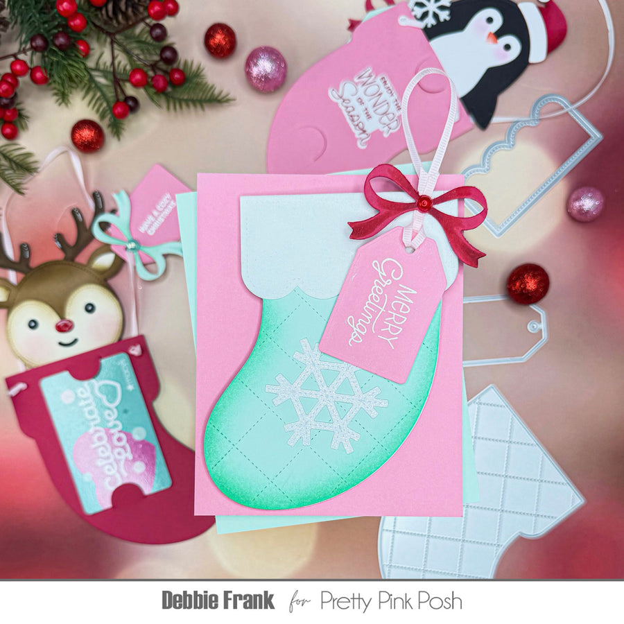 A Christmas stocking card craft design by Debbie Frank for Pretty Pink Posh, featuring a mint green stocking with a white snowflake on a pink background, adorned with a pink hanging tag that reads 'Merry Christmas' in white script. The card is styled with holiday decorations including a reindeer ornament, red berries, and a penguin craft piece on a soft pink background.