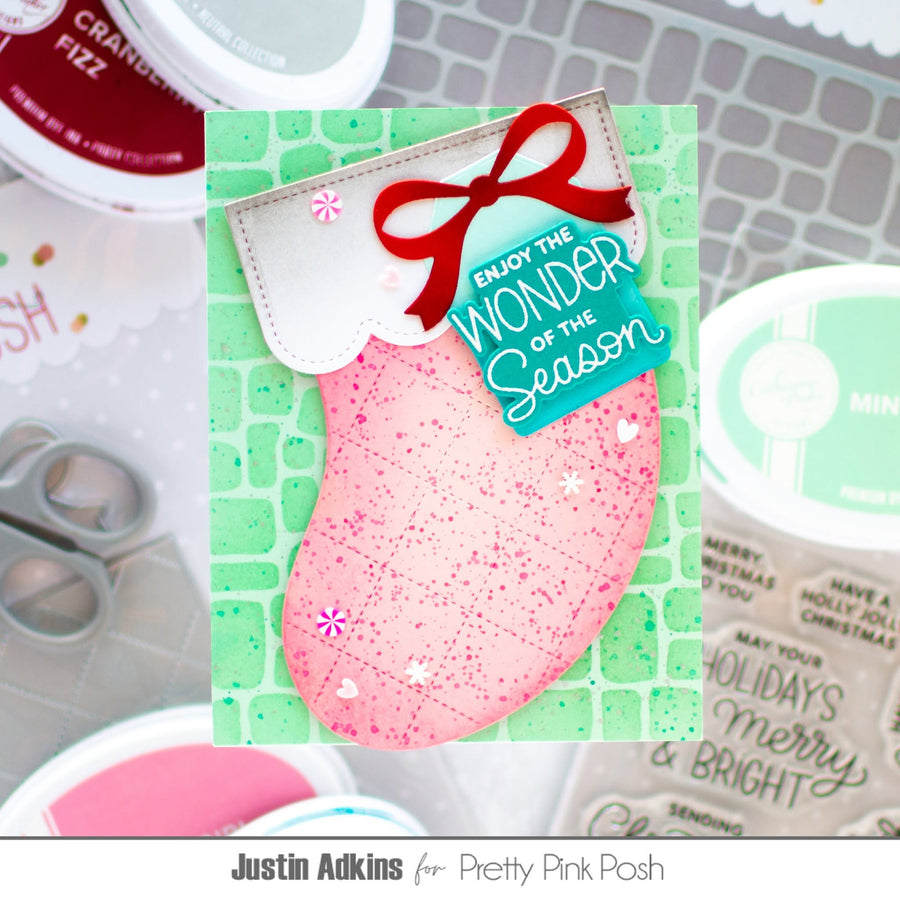 A handmade Christmas stocking card featuring a pink stocking with a red bow, a turquoise tag reading 'ENJOY THE WONDER OF The Season,' and a green patterned background. The card is displayed on a crafting mat with paint pots and scissors around it.