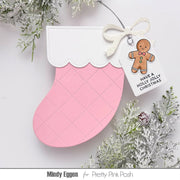 A pink and white Christmas stocking handmade greeting card or ornament with a stitched diamond pattern, decorated with a white glitter bow and a hanging tag that says 'HAVE A HOLLY JOLLY CHRISTMAS' with a gingerbread man illustration. The ornament is styled with frosted greenery on a white background, with 'Mindy Eggen for Pretty Pink Posh' printed at the bottom.