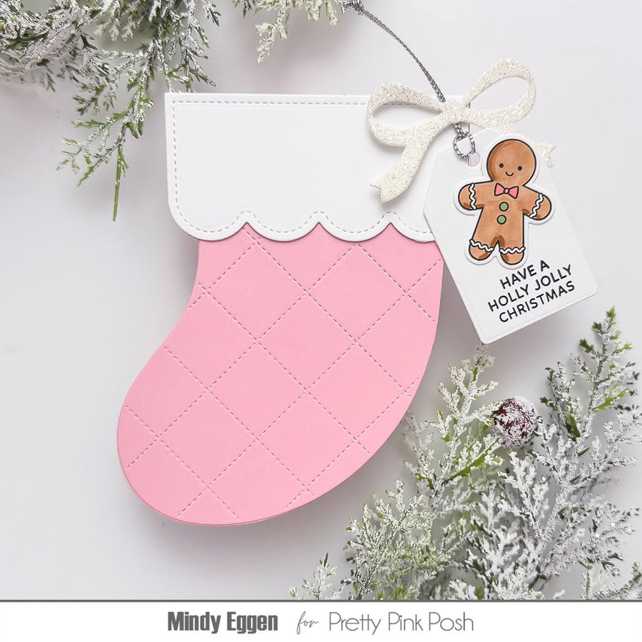 A pink and white Christmas stocking handmade greeting card or ornament with a stitched diamond pattern, decorated with a white glitter bow and a hanging tag that says 'HAVE A HOLLY JOLLY CHRISTMAS' with a gingerbread man illustration. The ornament is styled with frosted greenery on a white background, with 'Mindy Eggen for Pretty Pink Posh' printed at the bottom.