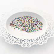 Multicolor craft sprinkles or craft beads scattered on a white plate with a decorative white paper doily underneath, set against a clean white background. Pretty Pink Posh Sugar Cookie Shaker Beads