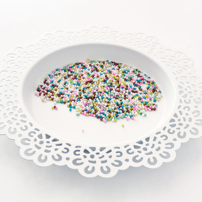 Multicolor craft sprinkles or craft beads scattered on a white plate with a decorative white paper doily underneath, set against a clean white background. Pretty Pink Posh Sugar Cookie Shaker Beads