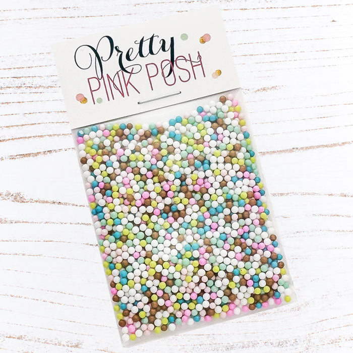 A clear plastic bag filled with many small pastel round craft sprinkles or beads, attached to a white header card that reads 'Pretty Pink Posh.' The package is photographed on a white wood surface. Pretty Pink Posh Sugar Cookie Shaker Beads