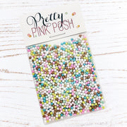 A clear plastic bag filled with many small pastel round craft sprinkles or beads, attached to a white header card that reads 'Pretty Pink Posh.' The package is photographed on a white wood surface. Pretty Pink Posh Sugar Cookie Shaker Beads