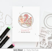 A white greeting card with a hand-drawn holiday illustration of gingerbread cookies, a candy cane, and a Christmas tree, with the words 'sweet HOLIDAY WISHES' below. The card is styled on a white background with craft markers, die-cut outlines, and another card featuring similar holiday line art.