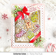 A handmade Christmas greeting card featuring gingerbread cookies, a stocking, candy canes, pine branches, and a red bow, displayed on a white textured surface with festive props. The card is branded for Catherine Cohen and Pretty Pink Posh.