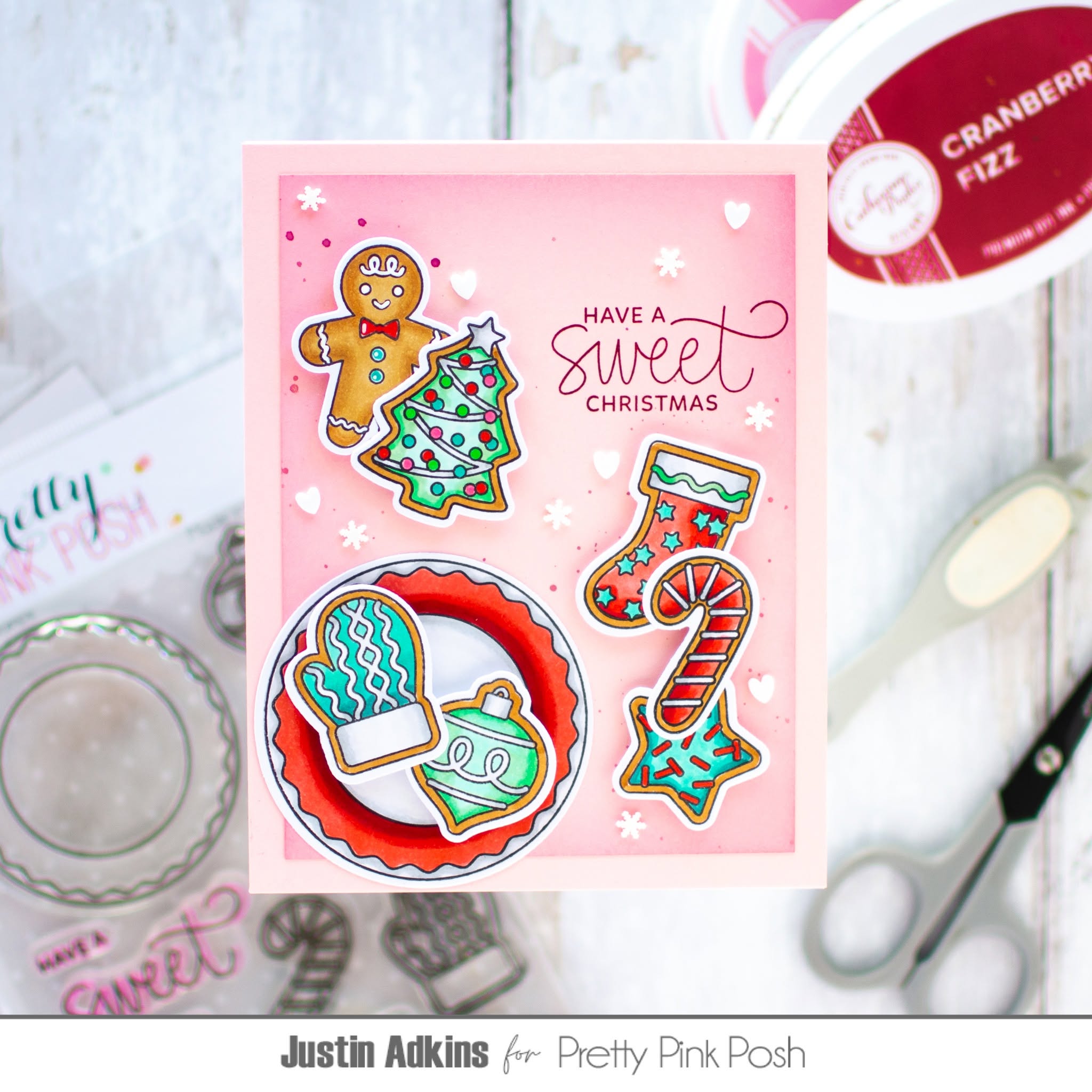 A handmade Christmas greeting card featuring gingerbread cookies, a tree, a stocking, and a mitten with the text 'HAVE A sweet CHRISTMAS' on a pink background, styled on a white wood surface with craft tools and Pretty Pink Posh branding visible.