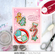 A handmade Christmas greeting card featuring gingerbread cookies, a tree, a stocking, and a mitten with the text 'HAVE A sweet CHRISTMAS' on a pink background, styled on a white wood surface with craft tools and Pretty Pink Posh branding visible.