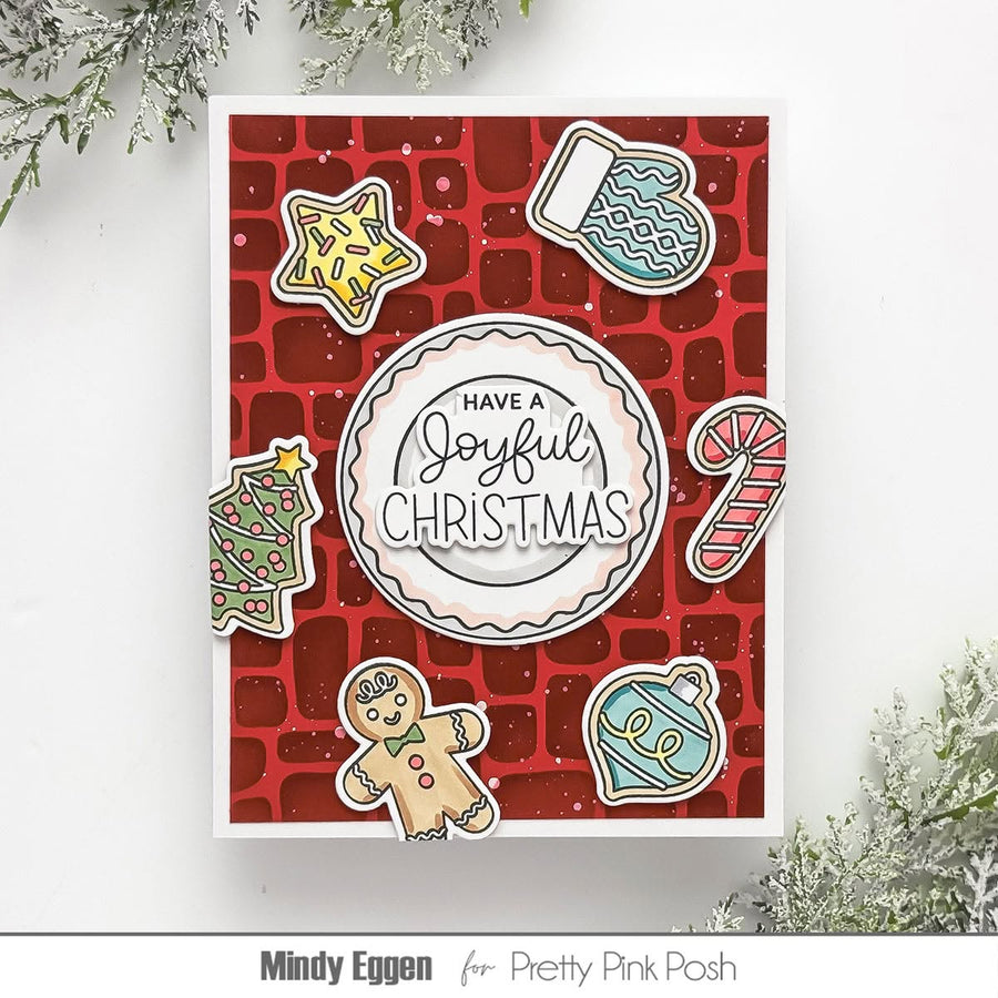 A handmade Christmas greeting card featuring a red brick pattern background, a central scalloped circle with the text 'HAVE A Joyful CHRISTMAS,' and festive cookie illustrations including a star, mitten, candy cane, ornament, gingerbread man, and tree. The card is photographed on a white surface with decorative greenery in the corners.