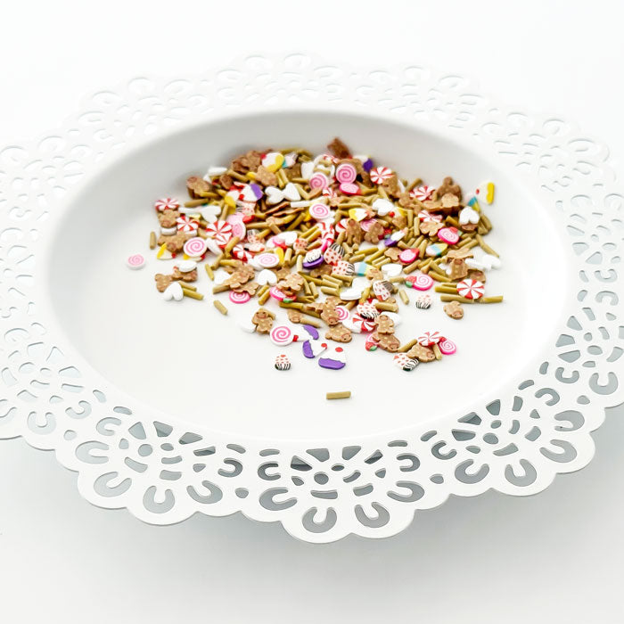 A white ceramic plate with a decorative cutout scalloped rim, filled with a colorful assortment of craft embellishments, beads, and sprinkles on a white background. Pretty Pink Posh Sweet Gingerbread Clay Confetti