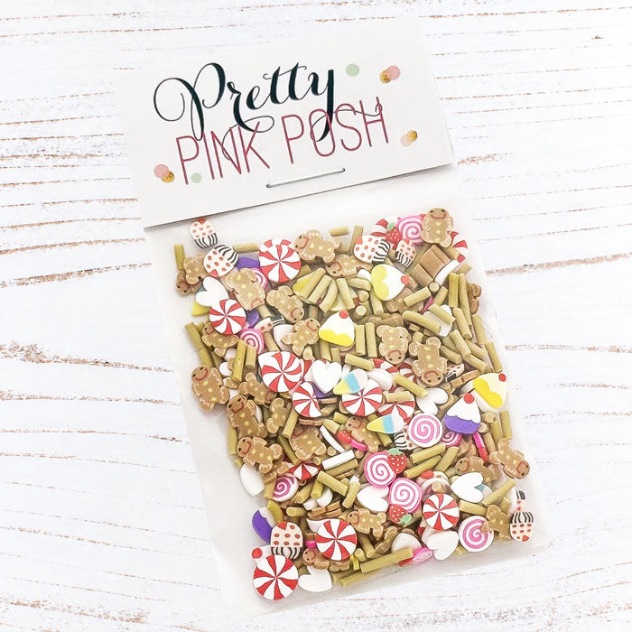 A clear plastic bag filled with decorative craft sprinkles and candy-shaped embellishments, sealed with a white header card that reads 'Pretty Pink Posh.' The product is photographed on a white wood surface. Pretty Pink Posh Sweet Gingerbread Clay Confetti