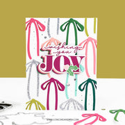A handmade layered greeting card with the words 'Wishing you JOY' in pink and white, decorated with colorful ribbon and pom-pom cutouts in pink, green, teal, and silver glitter. The card is displayed on a white surface against a mustard yellow background, with crafting tools visible at the bottom.