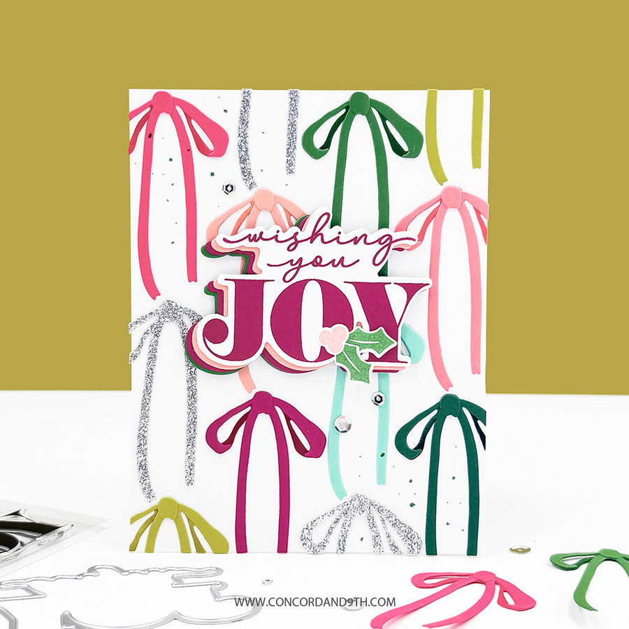 A handmade layered greeting card with the words 'Wishing you JOY' in pink and white, decorated with colorful ribbon and pom-pom cutouts in pink, green, teal, and silver glitter. The card is displayed on a white surface against a mustard yellow background, with crafting tools visible at the bottom.